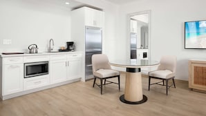Une cuisine moderne avec des armoires blanches, un réfrigérateur en acier inoxydable, un four encastré et une machine à café. On y trouve une table ronde avec deux chaises et un grand miroir au-dessus.