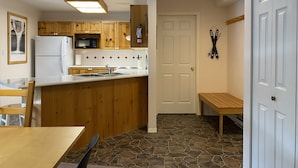 Een compacte keuken met houten kastjes, een witte koelkast, een magnetron en een spoelbak. Er is een bankje en een deur met een ski-houder aan de muur.