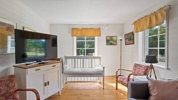 A living room with a television, a white bench, a wooden floor, and a window with curtains.