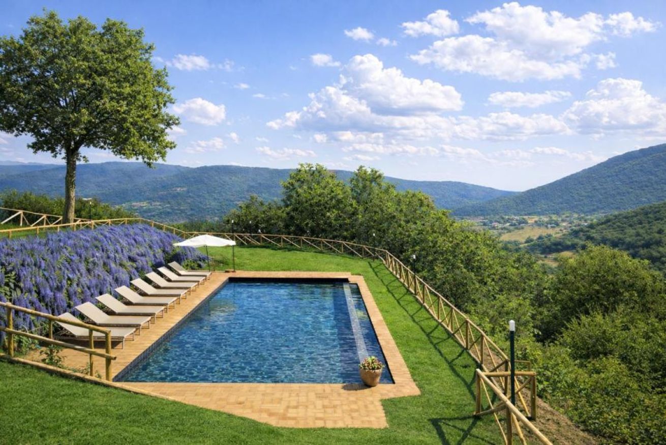 A rectangular swimming pool with a wooden deck and lounge chairs, surrounded by a wooden fence, set against a backdrop of green hills and a clear sky.