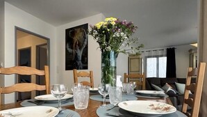Une table à manger est dressée avec des assiettes, des couverts et des verres, entourée de chaises en bois. Un vase rempli de fleurs est posé sur la table.