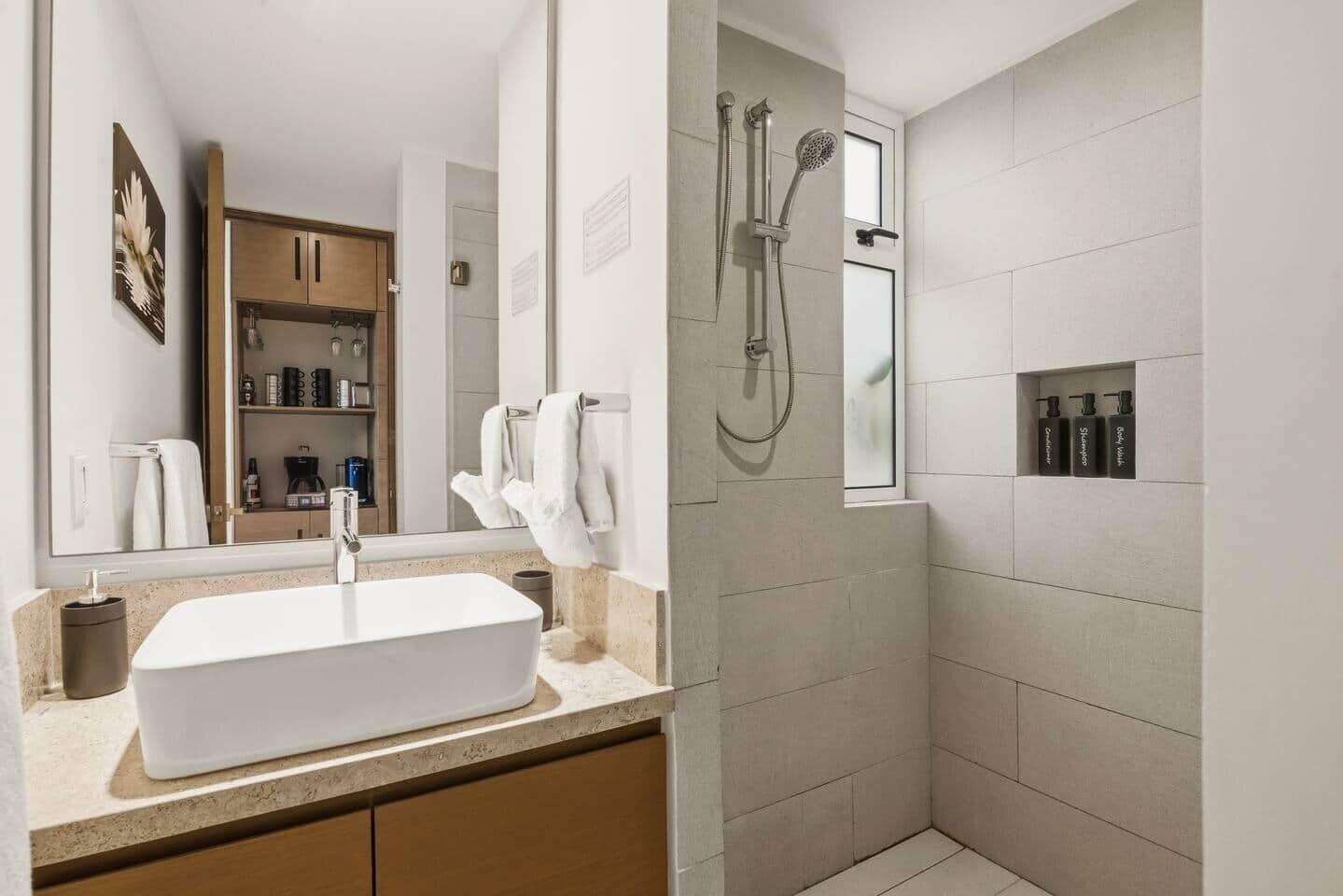 Een moderne badkamer met een grote spiegel, een rechthoekig wastafelmeubel, een douche met handdouche en een klein schapje met toiletartikelen.