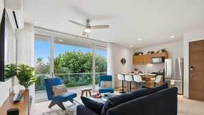 Een moderne woonkamer met een bank, fauteuils, een salontafel en een televisie. Er is een balkon met uitzicht op groen.