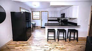 Eine kompakte Küche mit einem schwarzen Kühlschrank, einer Mikrowelle und einem Spülbecken. Drei Hocker und ein Esstisch mit Stühlen stehen bereit. Der Boden ist Holzboden, die Decke hat freiliegende Holzbalken.