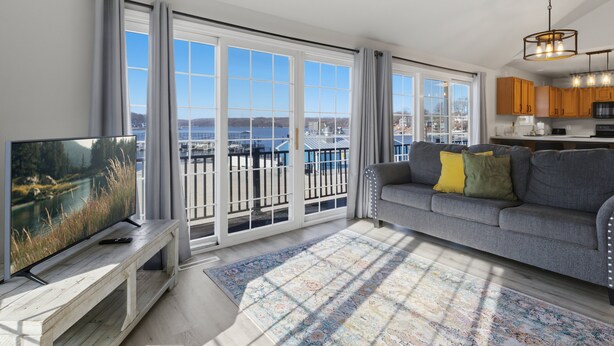 A modern living room with a large TV, a grey sofa with yellow pillows, a wooden bench, and a patterned rug. - Lakefront Home in Osage Beach 28mm at Miller's Landing (Osage Beach)