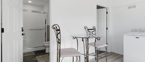 Een kleine, schone kamer met een witte deur, een glazen tafel, twee metalen stoelen, een wasmachine en een kamerkleed met patroon op de vloer.