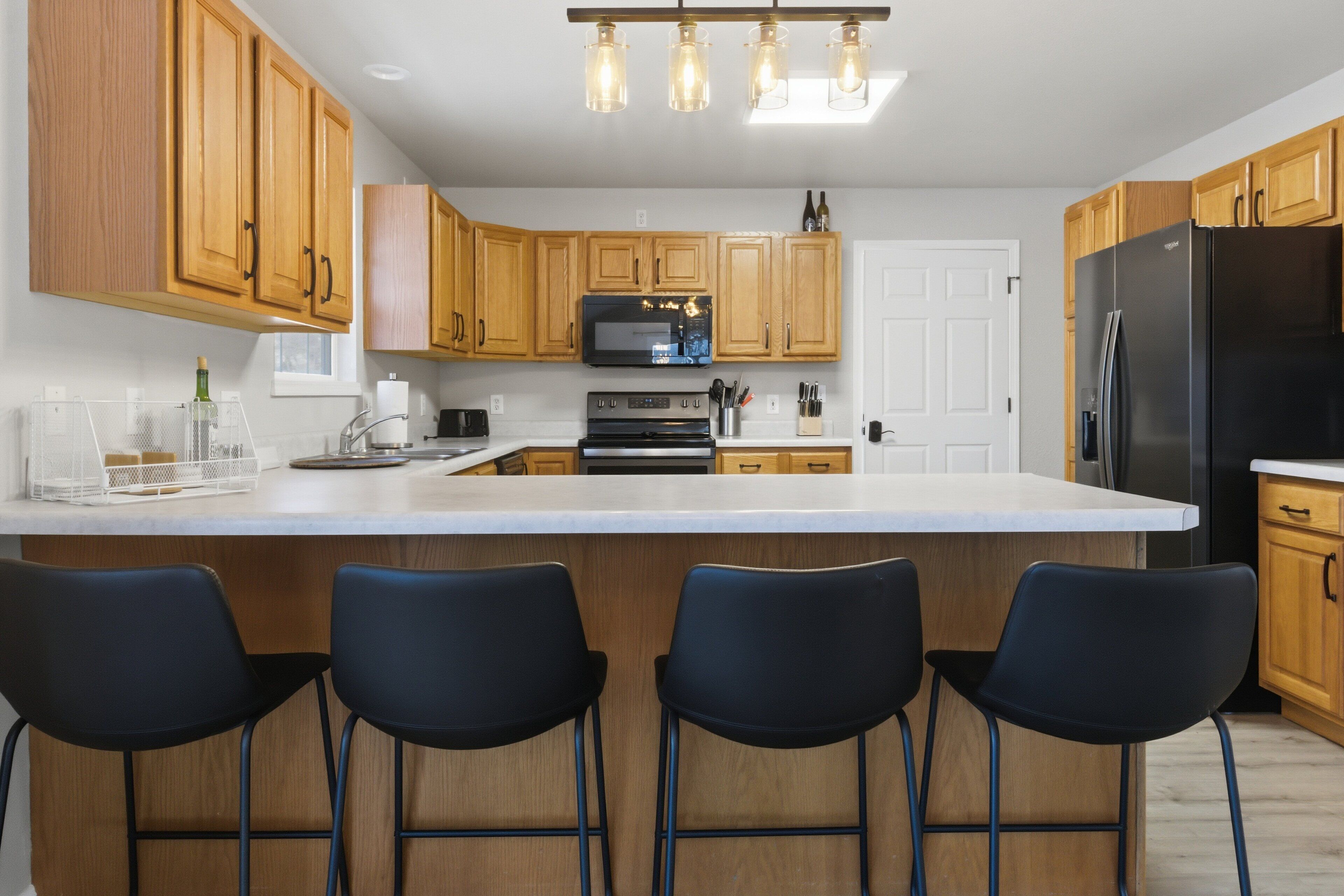 A modern kitchen with wooden cabinets, a large island with bar stools, a black refrigerator, and a built-in oven.