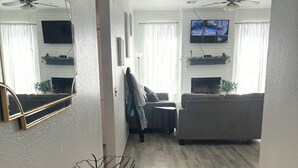 Un salon moderne avec un plancher en bois, une table à manger agrémentée d’un vase et d’une sculpture géométrique, un canapé, un téléviseur fixé au mur et un ventilateur de plafond.