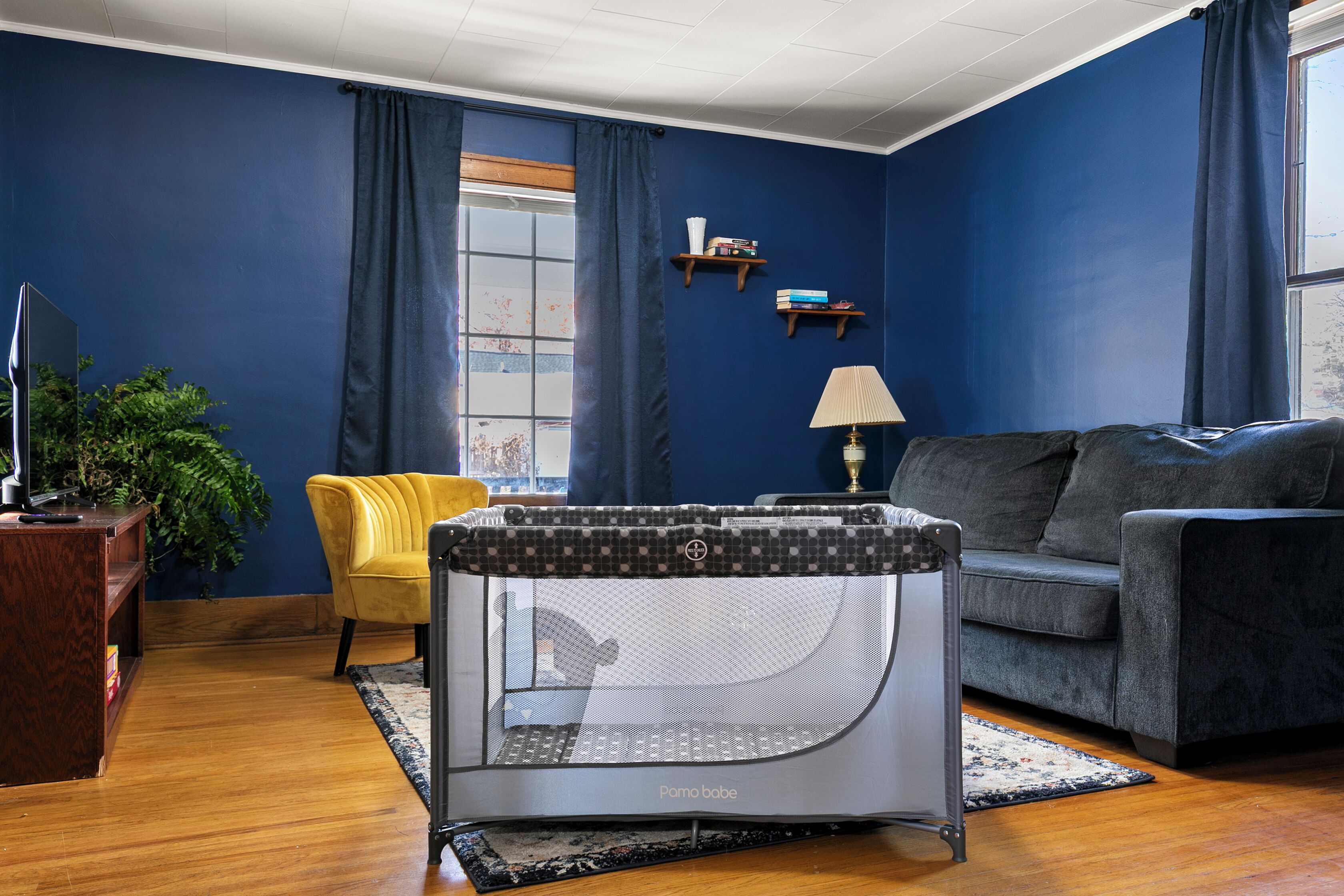 Una sala de estar con una pared azul, un sofá gris, una silla amarilla, un corral para bebés, un mueble para televisor y una planta.