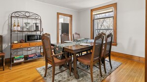A dining area with a wooden table, four chairs, a metal shelf with kitchen items, and a window with a view of trees.