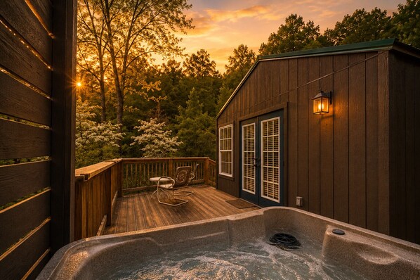 Une terrasse en bois avec un jacuzzi, un bâtiment en bois doté d’une porte, et une vue sur les arbres.