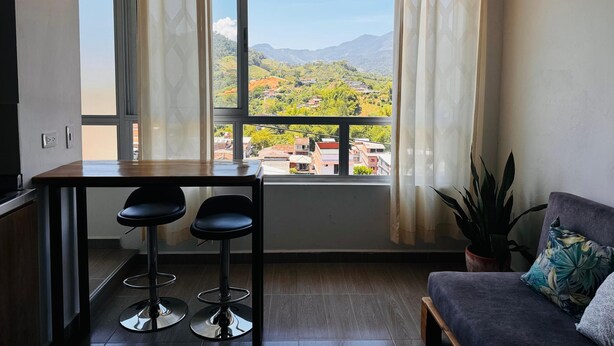 Una habitación con comedor, sofá y un ventanal con vistas a un paisaje verde y montañas.