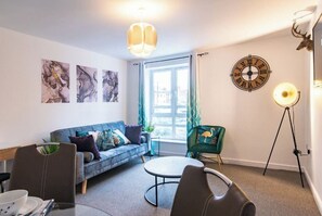 A modern living room with a sofa, a round table, and a chair. There is a wall clock and a lamp on a tripod stand. - Apartment - Sleeps 2 - Parking - City Centre Close (Coventry)