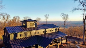 Eine zweistöckige Holzhütte mit einem Metall-Dach und zahlreichen Fenstern, eingebettet in eine bewaldete Landschaft.