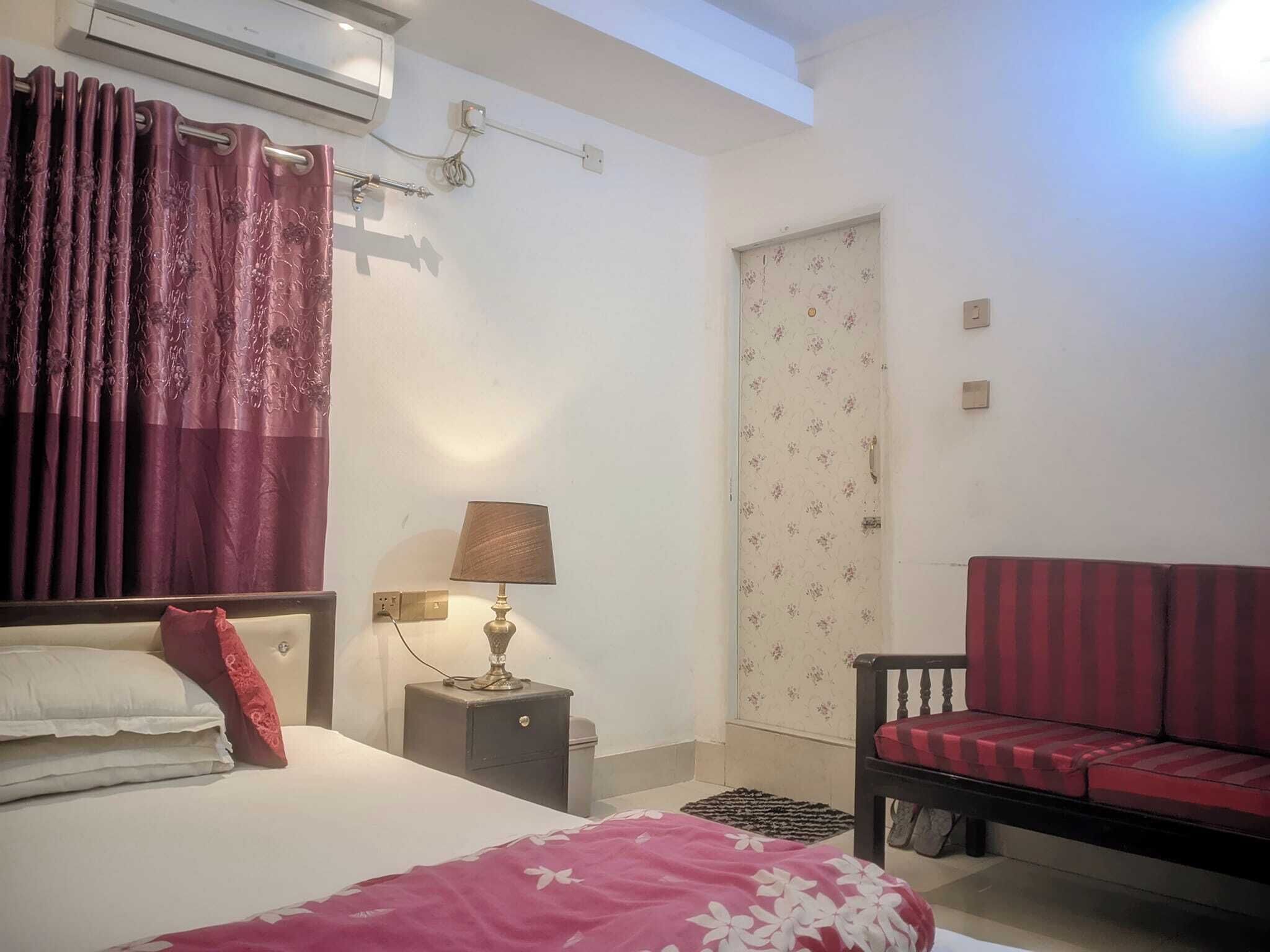 A bedroom with a bed, a nightstand with a lamp, a sofa, a door with floral wallpaper, and an air conditioning unit.