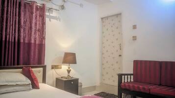 A bedroom with a bed, a nightstand with a lamp, a sofa, a door with floral wallpaper, and an air conditioning unit.