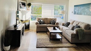 Ein Wohnzimmer mit einem beigen Sofa, zwei Kissen in Schwarz-Weiß-Muster, einem Holztisch und einem großen Fenster mit Blick auf Grünflächen.