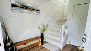 Eine Treppe führt zu einer Tür, an der ein Schild zum Ausziehen der Schuhe angebracht ist. Neben der Treppe befindet sich eine hölzerne Bank und ein Blumentopf mit Pflanzen auf dem Boden.