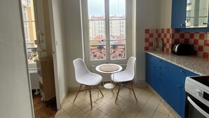 A kitchen with two white chairs, a round table, a hanging light fixture, a window with a view of buildings, and blue cabinets. - Bourgeois apartment 2 min from Bellecour. (Lyon)