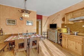 A dining area with a wooden table and chairs, a chandelier, and a kitchen with wooden cabinets and appliances. - Rochebrune 5th - Flat with Balcony for 4, Claviere, Italy (Claviere)