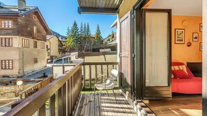 A wooden deck with a chair, a bed with red bedding, and a view of houses and trees. - Rochebrune 5th - Flat with Balcony for 4, Claviere, Italy (Claviere)