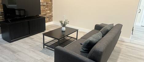 Una sala de estar moderna con televisor de pantalla plana, un sofá oscuro, una mesa de centro y una pared de ladrillo decorativa.