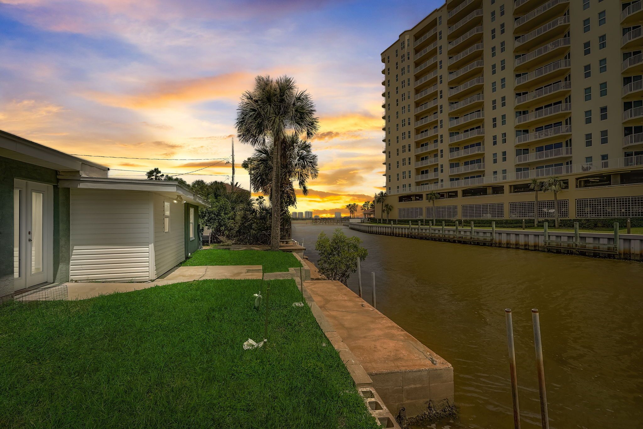 A riverside view with a tall building, a palm tree, and a grassy area.