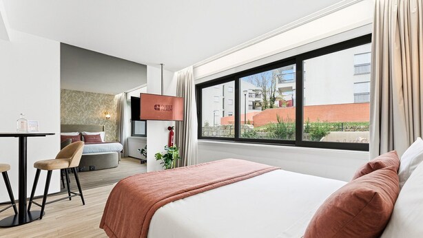 Habitación de hotel con una cama, un escritorio y una silla. Se ve una ventana con vistas a edificios.