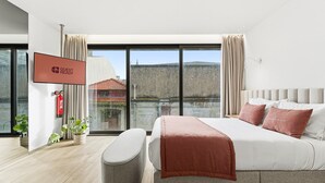 Ein modernes Hotelzimmer mit einem großen Bett, einem Flachbildfernseher, einem Feuerlöscher und Blick auf die Stadt durch große Fenster.