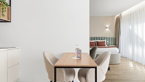 Una camera d'albergo moderna con un tavolo da pranzo in legno, due poltrone imbottite, un letto con testiera verde e un'ampia finestra con tende leggere.