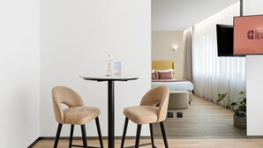 A modern hotel room with a bed, a TV mounted on the wall, a small round table with two chairs, and a bottle of water on the table. - GuestReady - Alma Invicta 'Casa da Música II' (Porto)