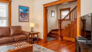 A living room with a brown leather sofa, a wooden coffee table, a framed picture, a lamp, and a staircase leading to another room. - The Osborne House 1930 near Gore Mountain with ARCADE! Air conditioned! (North Creek)