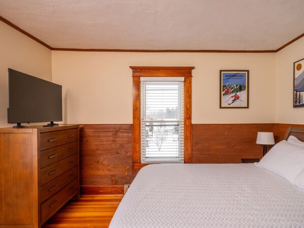 Une chambre à coucher avec une commode en bois, un lit avec une literie blanche, une fenêtre avec des stores et un tableau encadré au mur.