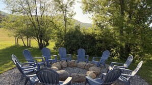 A circular seating arrangement with Adirondack chairs around a central fire pit, set on a gravel surface with a grassy field and trees in the background. - The Osborne House 1930 near Gore Mountain with ARCADE! Air conditioned! (North Creek)