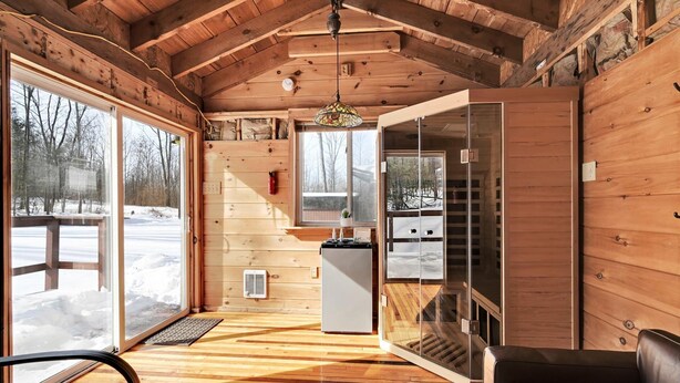 L’intérieur chaleureux d’un chalet avec des murs en bois, une porte vitrée donnant sur l’extérieur, un petit coin cuisine et un espace salon avec un canapé en cuir.