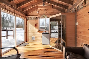 Interior acogedor de una cabaña con paredes de madera, un ventanal que da al exterior, una pequeña cocina y una zona de estar con un sofá de cuero.