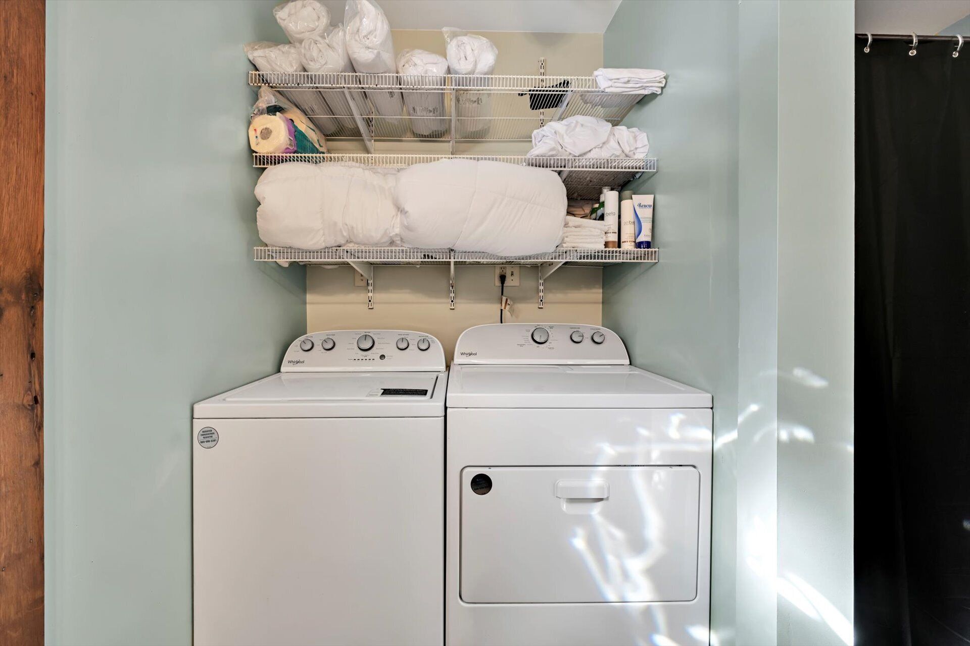 Uma área de lavanderia com duas máquinas de lavar e secar Whirlpool, uma prateleira de metal acima com toalhas dobradas e produtos de lavagem.
