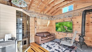 A cozy cabin interior with a wooden ceiling, a glass door leading outside, a brown leather sofa, a television, a fireplace, and a patterned rug.