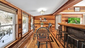 Un intérieur de chalet chaleureux avec des murs et des planchers en bois, un coin repas avec une table et des chaises, un bar avec des tabourets et un téléviseur fixé au mur.