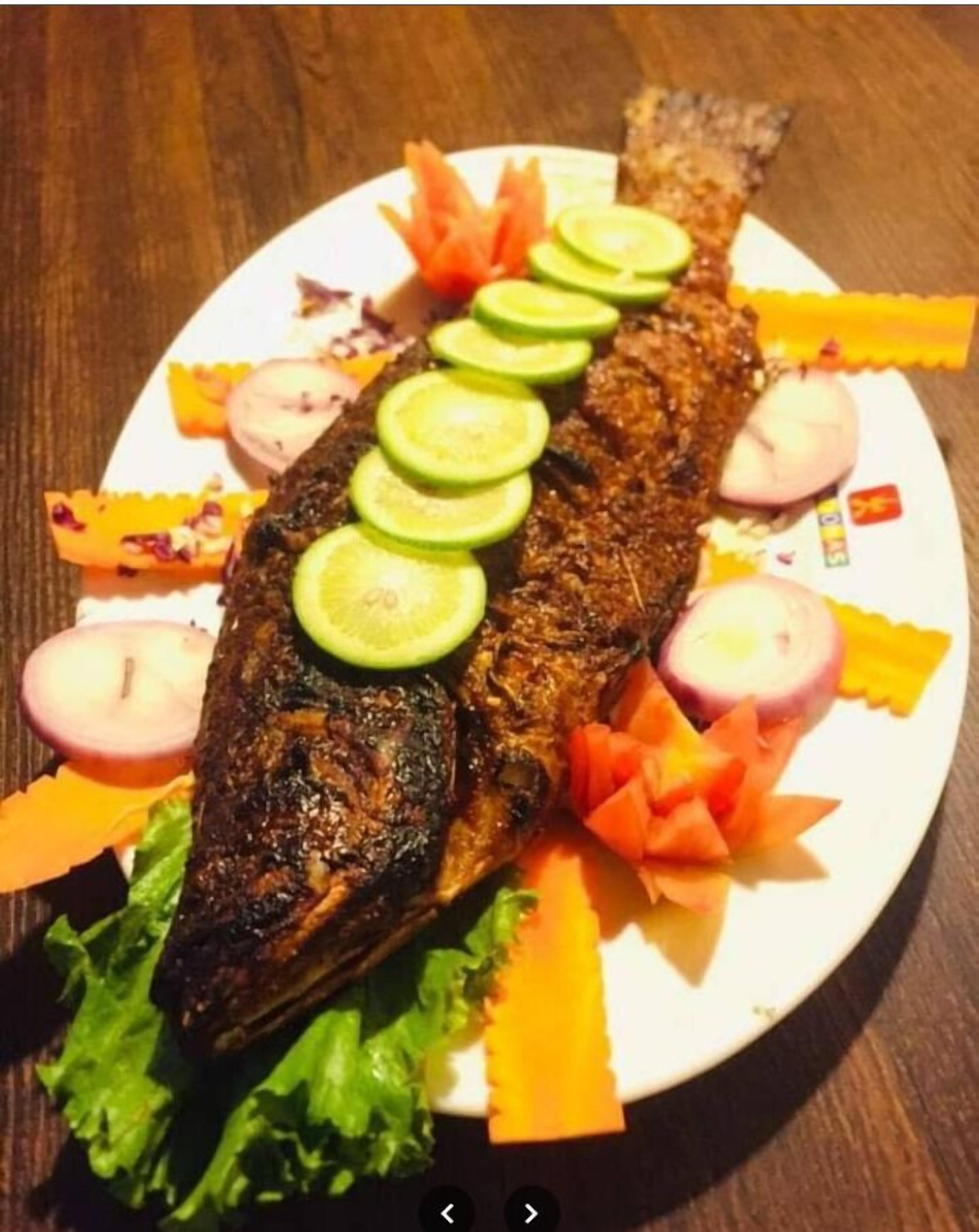 A whole grilled fish on a white plate with cucumber slices, carrot slices, and onion rings.