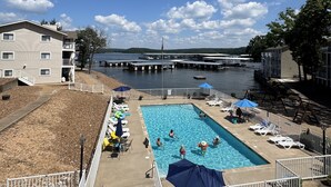 A swimming pool with lounge chairs and umbrellas, a dock, and a building with a balcony overlooking the water.