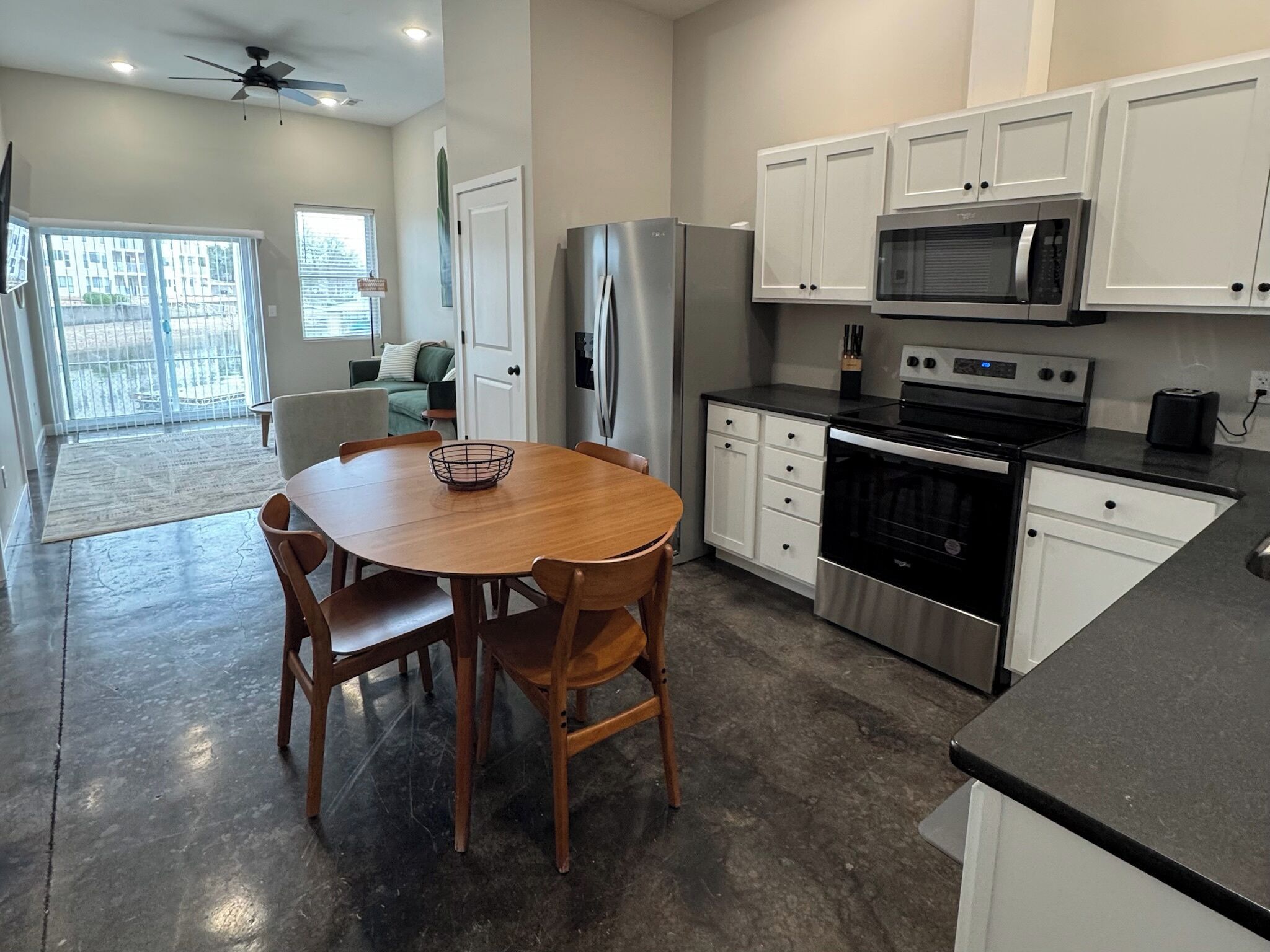 A modern kitchen with white cabinets, a stainless steel refrigerator, a microwave, and an oven. There is a dining table with chairs and a ceiling fan.