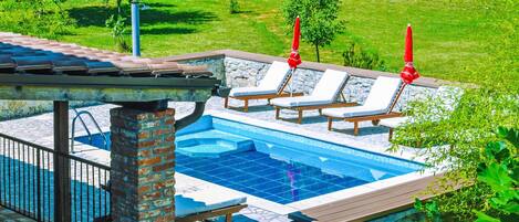 Una piscina con sillas de descanso y sombrillas, rodeada de muros de piedra y vegetación.