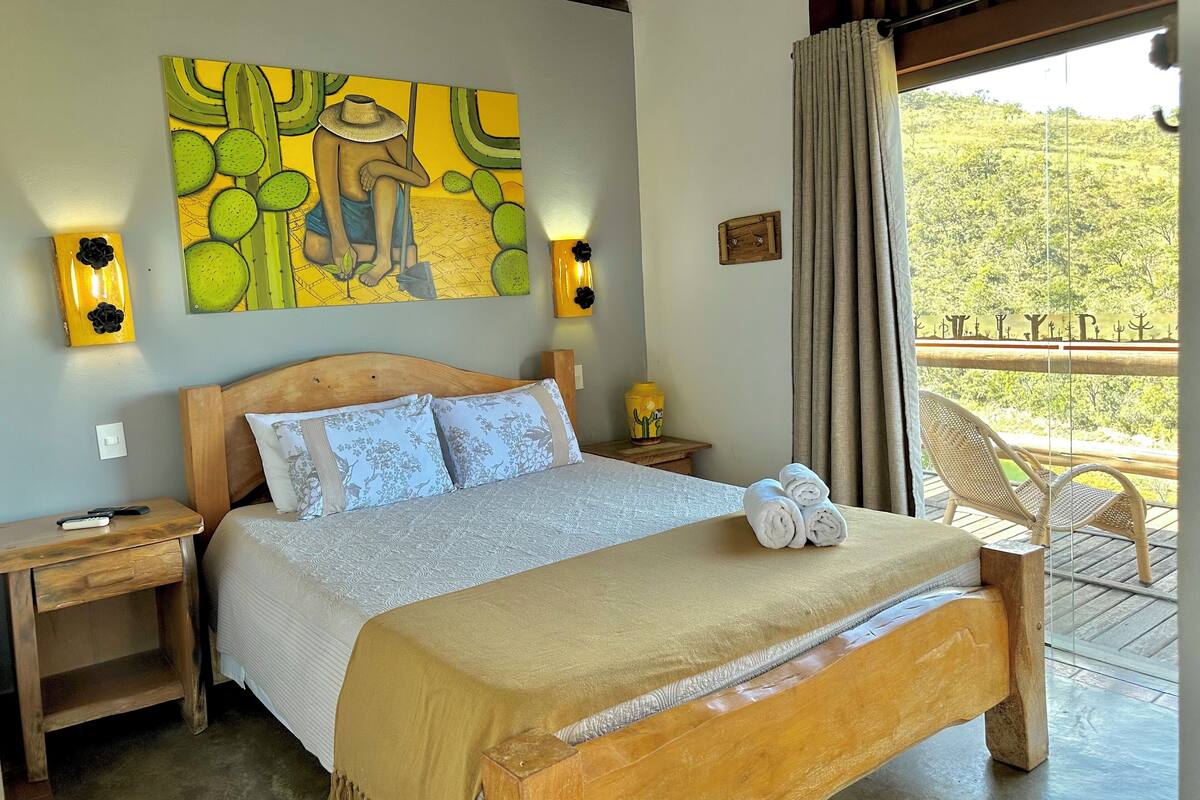 A bedroom with a wooden bed, a large painting, two bedside tables with lamps, and a view of greenery through a window.