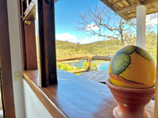 A view from a window with a wooden ledge, a terracotta pot with a painted object, and a landscape with a river and hills. - Pousada Fecho da Serra (Capitólio)