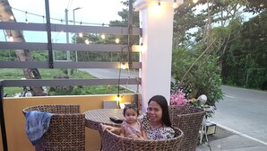 A seating area with wicker chairs and a table, a white pillar with lights, and a view of a road and greenery.