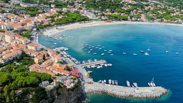 Vista aerea di una località costiera con un porto turistico, edifici con tetti in tegole rosse e un mare cristallino.