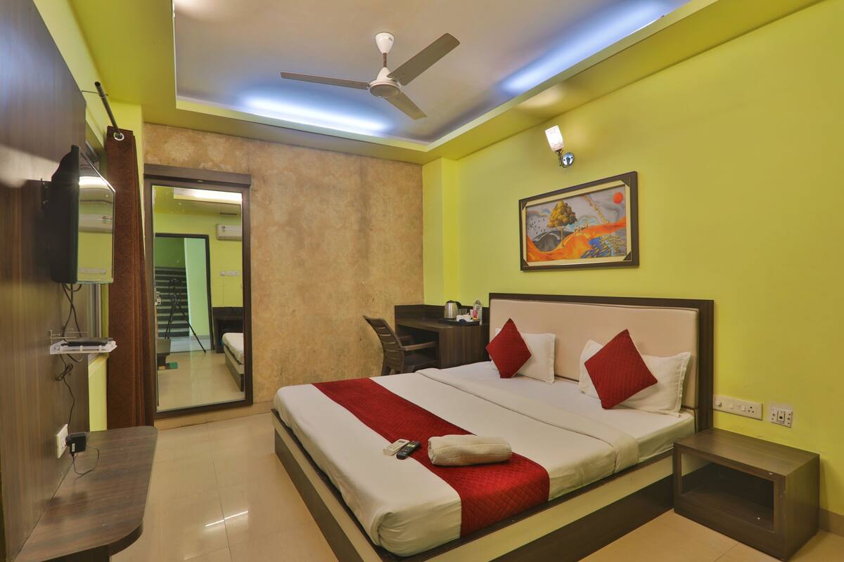 A hotel room with a bed, a desk, a chair, a ceiling fan, a painting, and a mirror.
