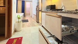 A modern kitchen with white cabinets, a built-in oven, and a microwave. There is a red welcome mat at the entrance. - Trovero Suites (Greater Noida)