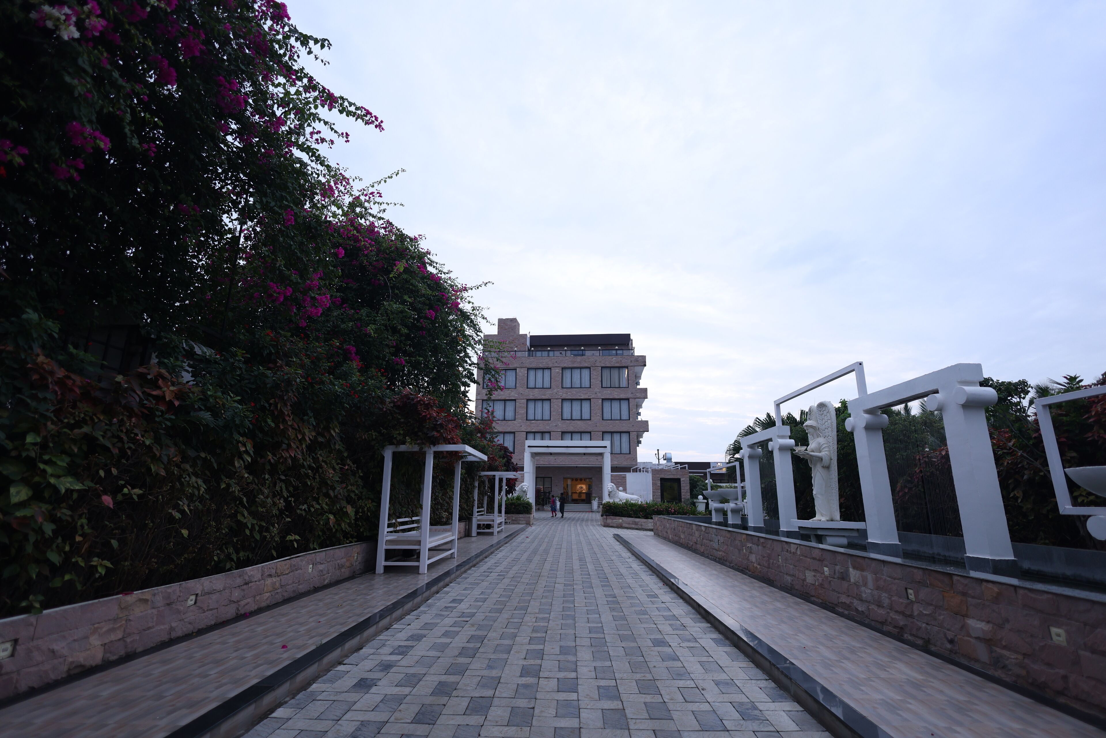 A paved pathway leading to a building with a modern architectural style, surrounded by greenery and white decorative elements.